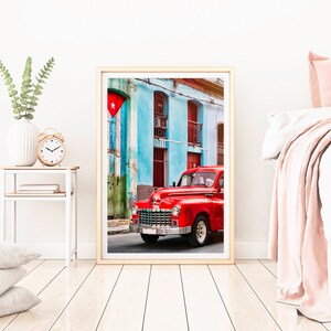 May include: A vintage red car parked in front of a colorful building with a Cuban flag hanging from the wall. The building has blue walls, red doors, and white trim.