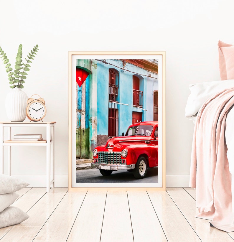 May include: A vintage red car parked in front of a colorful building with a Cuban flag hanging from the wall. The building has blue walls, red doors, and white trim.