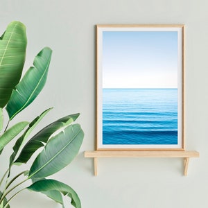 May include: A framed photograph of a blue ocean with a light blue sky above. The photograph is on a wooden shelf against a light green wall.