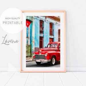 May include: A red vintage car parked in front of a blue and green building with a Cuban flag hanging on the side. The car is in focus and the building is out of focus.
