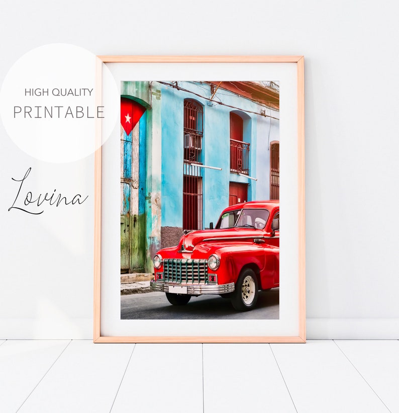 May include: A red vintage car parked in front of a blue and green building with a Cuban flag hanging on the side. The car is in focus and the building is out of focus.