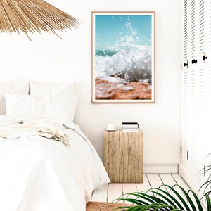 May include: A framed print depicting a crashing wave. The image shows the moment the wave breaks, with white water against orange-brown rocks. The sky is a clear, light blue. The print is displayed in a light-coloured wooden frame.