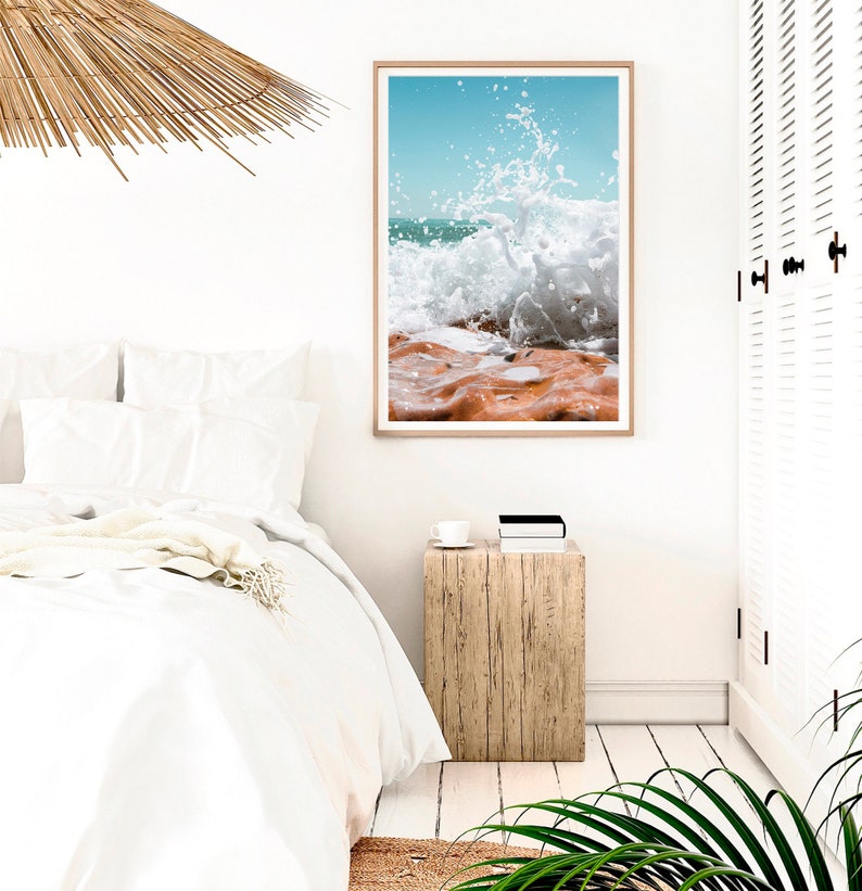 May include: A framed print depicting a crashing wave. The image shows the moment the wave breaks, with white water against orange-brown rocks. The sky is a clear, light blue. The print is displayed in a light-coloured wooden frame.