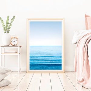 May include: A framed print of a blue ocean with waves and a light blue sky. The print is hanging on a white wall in a bedroom setting.