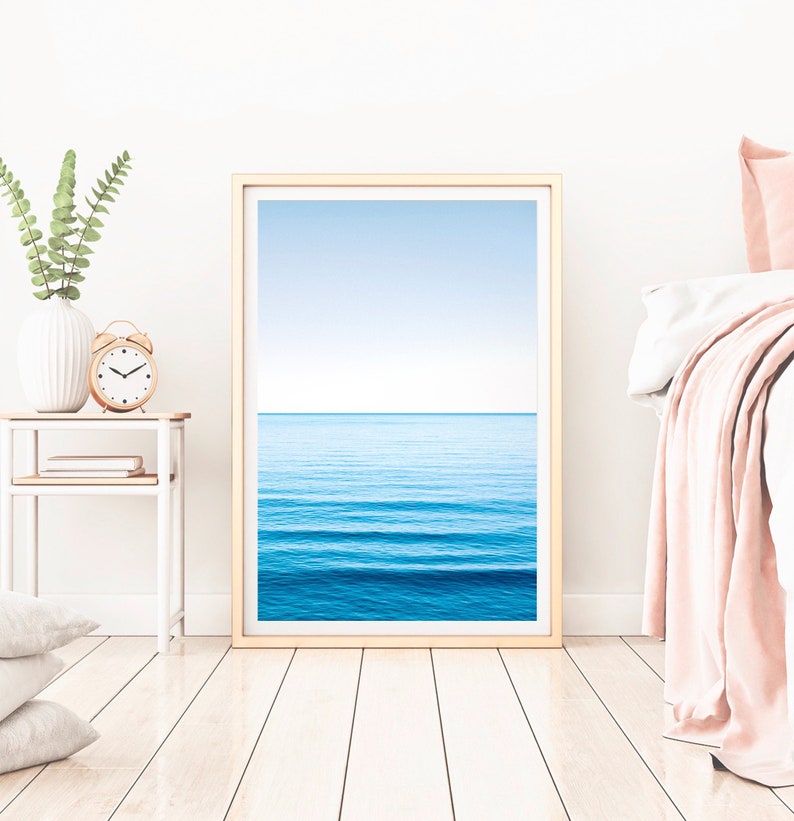May include: A framed print of a blue ocean with waves and a light blue sky. The print is hanging on a white wall in a bedroom setting.