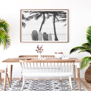 K&ouml;nnte beinhalten: Schwarzwei&szlig;foto von Surfbrettern, die an einer Palme an einem Strand lehnen. Das Foto ist in einem hellen Holzrahmen gerahmt und h&auml;ngt an einer wei&szlig;en Wand &uuml;ber einem Esstisch aus Holz mit wei&szlig;en St&uuml;hlen.