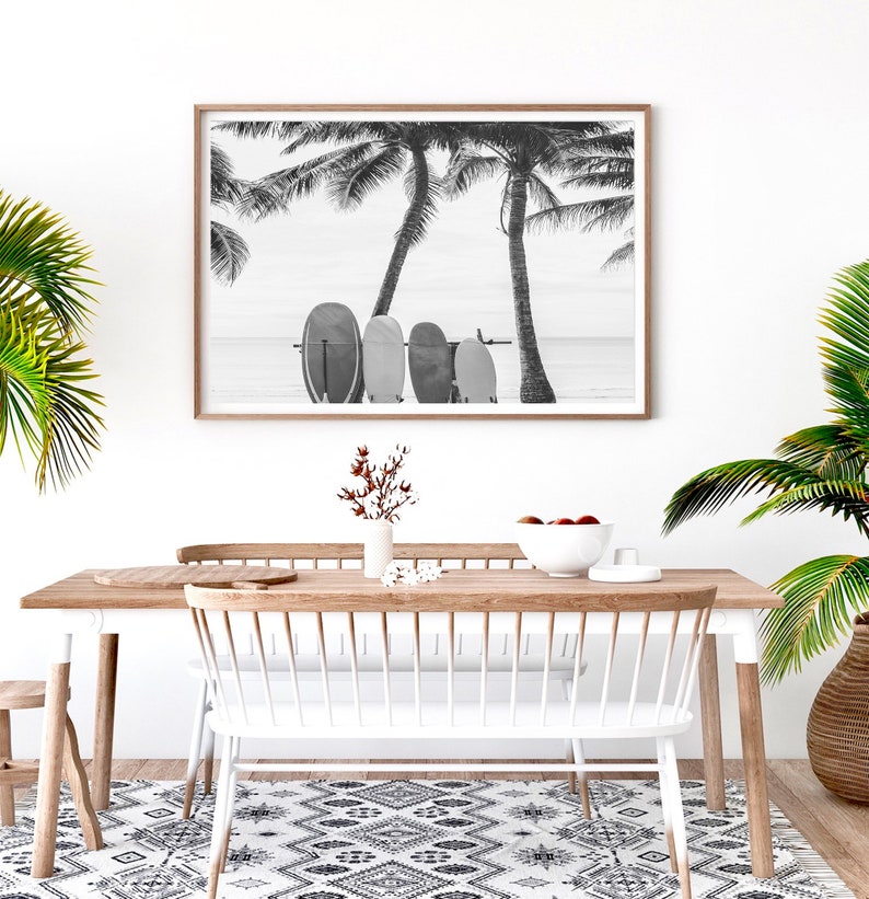 K&ouml;nnte beinhalten: Schwarzwei&szlig;foto von Surfbrettern, die an einer Palme an einem Strand lehnen. Das Foto ist in einem hellen Holzrahmen gerahmt und h&auml;ngt an einer wei&szlig;en Wand &uuml;ber einem Esstisch aus Holz mit wei&szlig;en St&uuml;hlen.