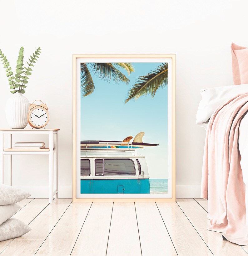 May include: A framed print of a blue vintage van with surfboards on the roof, parked on a beach with palm trees in the background.