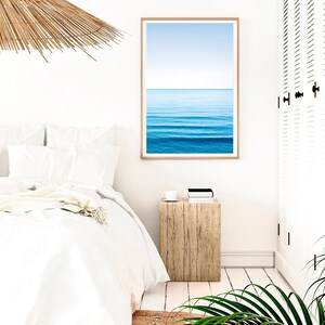 May include: A white bed with white bedding and a throw blanket. A wooden nightstand with a cup and a book on top. A framed print of a blue ocean with white waves and a blue sky above. A white door with louvered panels is in the background.
