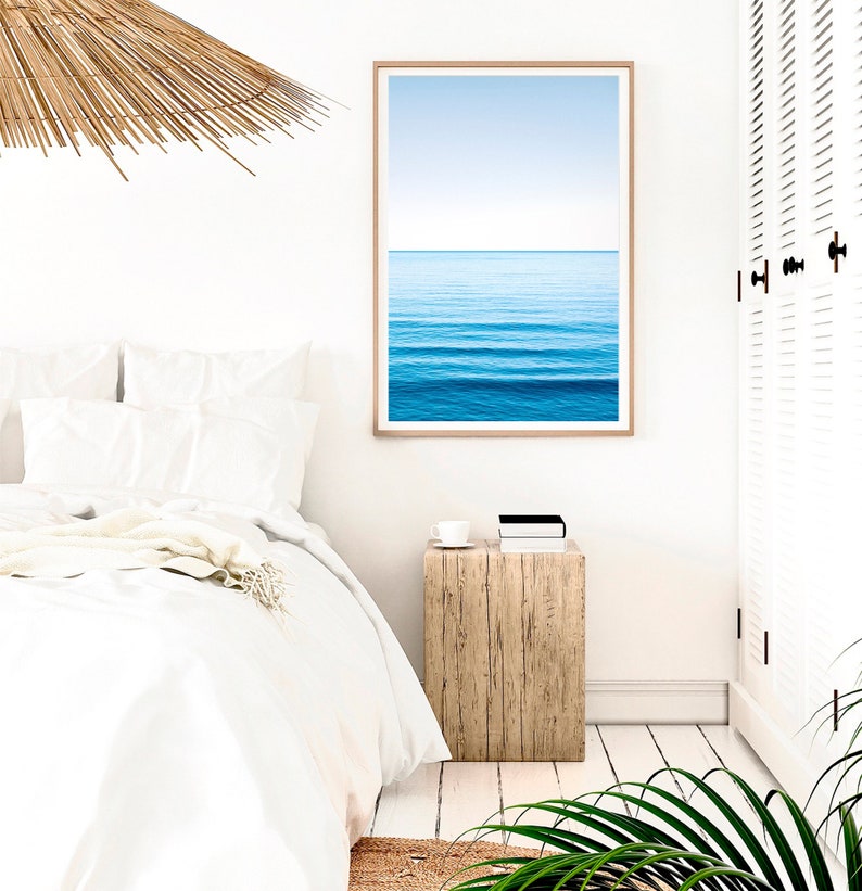 May include: A white bed with white bedding and a throw blanket. A wooden nightstand with a cup and a book on top. A framed print of a blue ocean with white waves and a blue sky above. A white door with louvered panels is in the background.