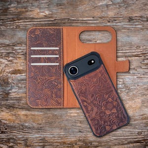 May include: A brown leather phone case with a floral embossed design. The case is open, revealing a brown interior and slots for cards. A black phone with a camera lens is inside the case. The case is on a wooden surface.