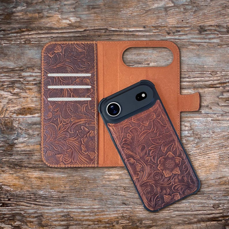 May include: A brown leather phone case with a floral embossed design. The case is open, revealing a brown interior and slots for cards. A black phone with a camera lens is inside the case. The case is on a wooden surface.