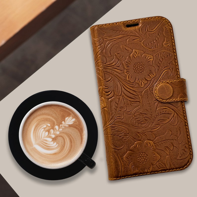 May include: A brown leather phone case with an embossed floral design. The case has a magnetic closure and a button clasp. A cup of latte with latte art sits on a black coaster next to the case.