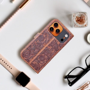 May include: Brown leather phone case with a floral embossed design. The case is open, revealing the phone's camera and screen. A smartwatch, glasses, and office supplies are arranged around the case.