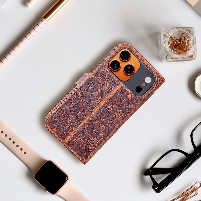 May include: Brown leather phone case with a floral embossed design. The case is open, revealing the phone's camera and screen. A smartwatch, glasses, and office supplies are arranged around the case.