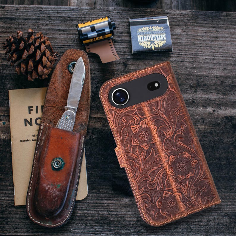 May include: A brown leather phone case with a floral embossed design, a pocket knife in a leather sheath, a pine cone, a film roll, and a matchbox are arranged on a weathered wooden surface. The phone case has a camera cutout.