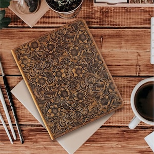Pode incluir: Um diário de couro marrom com um design floral em relevo. O diário está fechado e sobre uma superfície de madeira.
