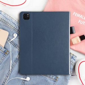 May include: A dark blue leather case for a tablet or phone. The case has a flap that covers the screen and a camera cutout. The case is lying on a white surface with a pair of jeans, a pink shirt, and a pair of white trainers.