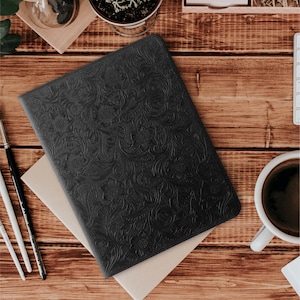Op de afbeelding: Een zwart lederen notitieboek met een reliëfpatroon van bloemen. Het notitieboek ligt op een houten oppervlak met een notitieblok en een kopje koffie.