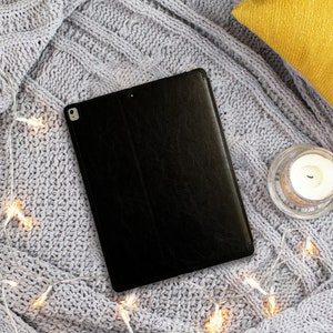 May include: A black leather tablet case on a grey knitted blanket with fairy lights.