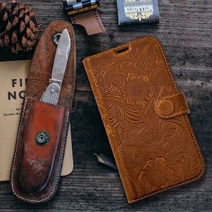 May include: A brown leather phone case with a floral embossed design and a snap closure. Beside it is a vintage pocket knife in a brown leather sheath. A pine cone, film canister, and matchbox are also present.