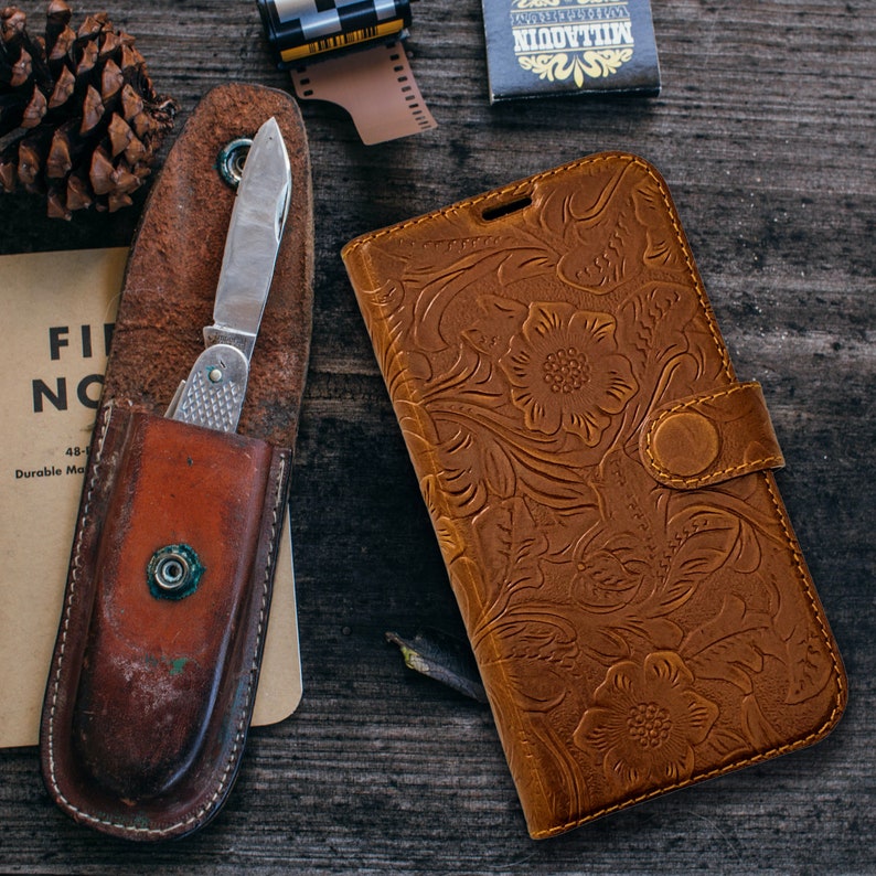 May include: A brown leather phone case with a floral embossed design and a snap closure. Beside it is a vintage pocket knife in a brown leather sheath. A pine cone, film canister, and matchbox are also present.