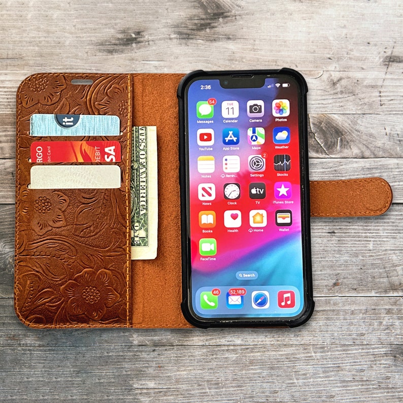 May include: Brown leather phone case with a floral embossed design. The case is open, revealing a smartphone with a black case, credit cards, and a dollar bill. The phone displays app icons on the screen.