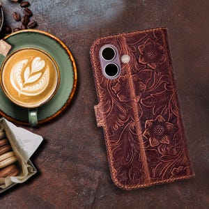 May include: A brown leather phone case with an embossed floral design. The case has a magnetic closure and a camera cutout. The background includes a cup of coffee with latte art, coffee beans, and pastries.