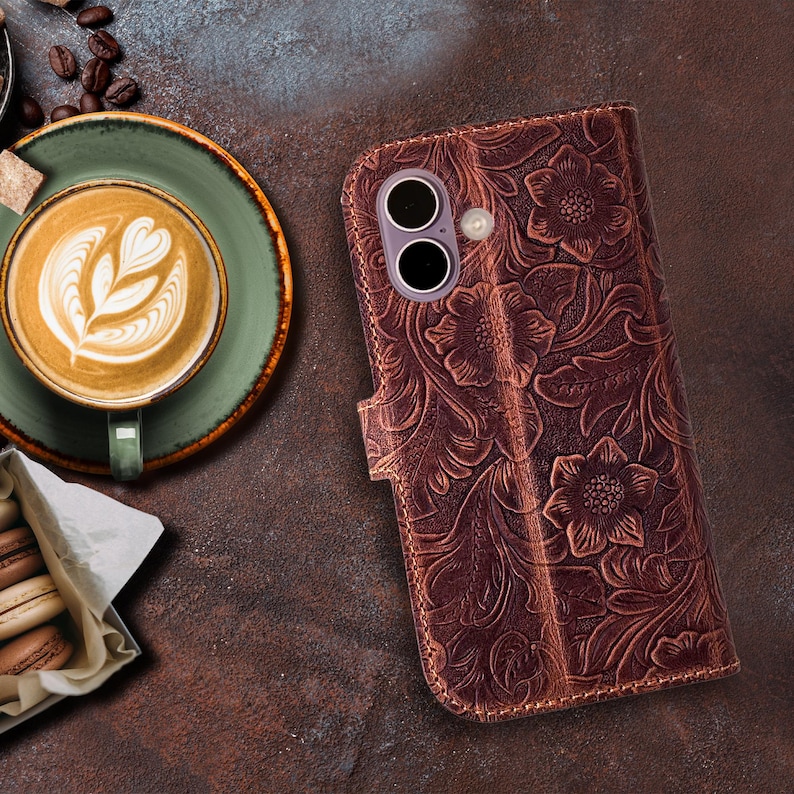 May include: A brown leather phone case with an embossed floral design. The case has a magnetic closure and a camera cutout. The background includes a cup of coffee with latte art, coffee beans, and pastries.