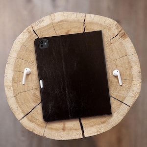 May include: A black leather case for a tablet or iPad. The case has a textured surface and a camera cutout. The case is lying on a wooden surface.