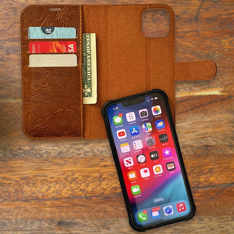 May include: A brown leather phone case with a floral pattern. The case has a flap that closes with a magnetic clasp. The case also has a slot for credit cards and cash. An iPhone is in the case, showing the home screen with various apps.