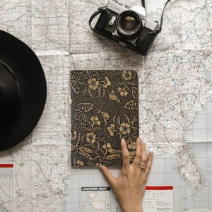 May include: A black leather journal with gold embossed floral and dragonfly designs. The journal is open and a hand is resting on the cover. A vintage camera and a black hat are also in the image.