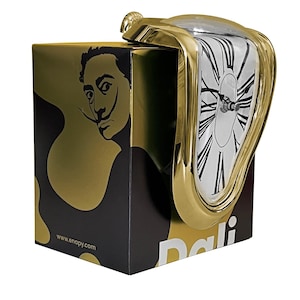 May include: A gold-colored, melting clock with a white clock face and black Roman numerals. The clock is on top of a black and gold box featuring a portrait of Salvador Dalí. The website address www.enapy.com is visible on the box.