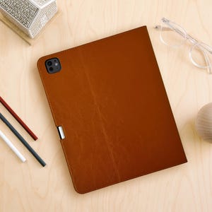 May include: A brown leather tablet case with a camera cutout and a silver clasp. The case is rectangular and has a stitched border. Other items on the wooden surface include pencils, glasses, and a decorative box.