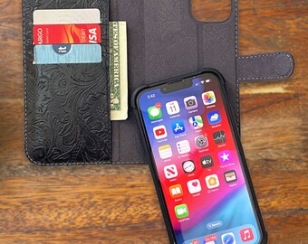 May include: A black leather phone case with a floral pattern and a built-in wallet. The case has a slot for credit cards and a clear plastic window for displaying a photo ID. The case is shown with a black iPhone with a bright screen displaying various apps.