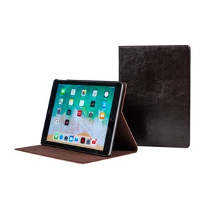 May include: A brown leather case for an iPad with a stand feature. The case is closed and the iPad is visible with the screen on, showing the home screen with various apps.