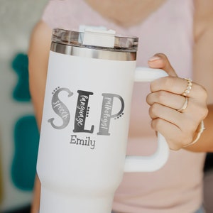 May include: White travel mug with a black and white design that says "SLP" with the words "speech", "language", and "pathologist" written vertically around the letters. The mug also has the name "Emily" written below the design.