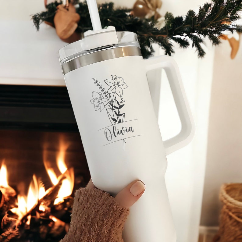 May include: White stainless steel travel mug with a black floral design and the name "Olivia" printed on it. The mug has a handle and a lid with a straw.