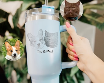 May include: A light blue stainless steel travel mug with a black and white engraved image of a dog and a cat. The mug has a handle and a lid with a straw. The text "Rio & Med" is engraved below the images.