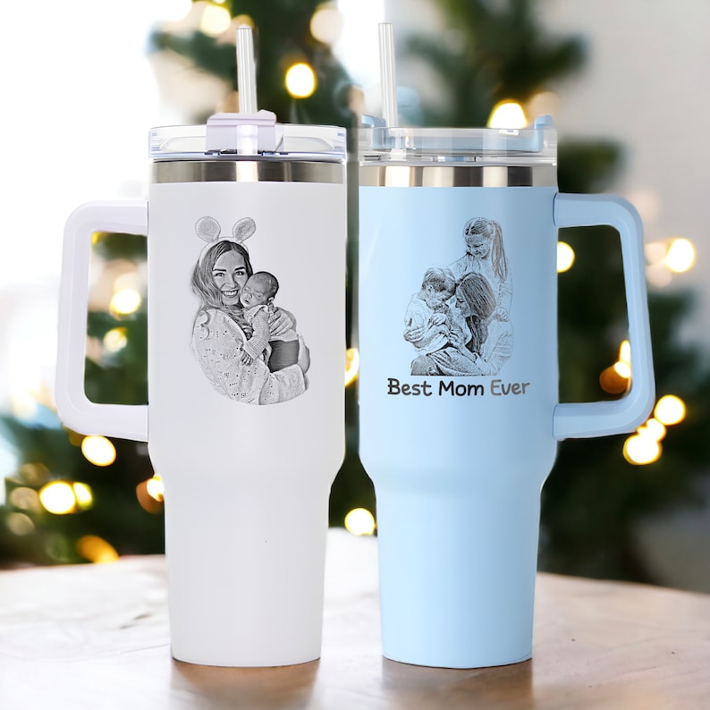 May include: Two stainless steel travel mugs with handles and lids. One mug is white with a black and white photo of a woman holding a baby. The other mug is light blue with a black and white photo of a woman and two children. The light blue mug has the text "Best Mom Ever" engraved on it.