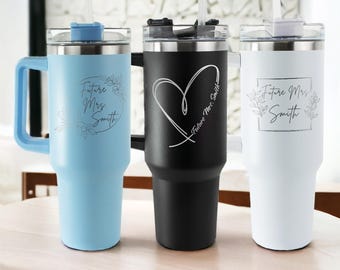 May include: Three stainless steel travel mugs with lids and straws. The mugs are engraved with different designs: "Future Mrs. Smith" with floral accents, a heart with "Future Mrs. Smith", and a square with "Future Mrs. Smith". The mugs are blue, black, and white.