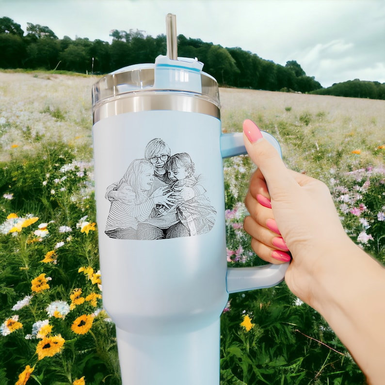 May include: A light blue stainless steel travel mug with a black and white photo of three people hugging. The mug has a lid with a straw and a handle.