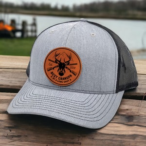 May include: A gray and black trucker hat with a brown leather patch that says "Best Grandpa Est. 2024 By A Long Shot" with a deer and crossed rifles.