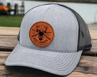 Op de afbeelding: Een grijze en zwarte trucker-pet met een bruin leren patch waarop staat "Best Grandpa Est. 2024 By A Long Shot" met een hert en gekruiste geweren.