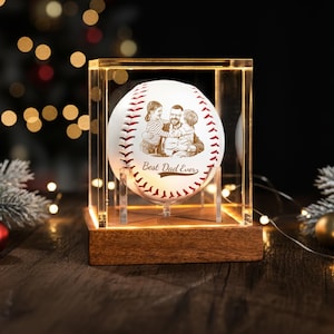 May include: A personalized baseball with a family portrait and the text "Best Dad Ever" displayed in a clear acrylic case with a wooden base. The baseball has red stitching and is illuminated by warm lighting.