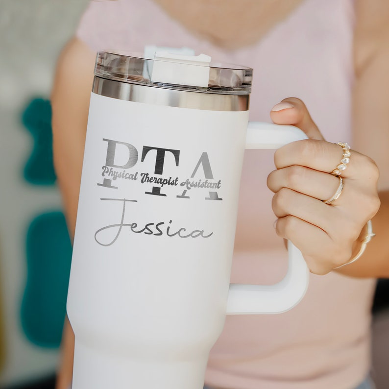 Pu&ograve; includere: Tazza da viaggio in acciaio inox bianco con manico nero. La tazza ha la scritta "PTA Physical Therapist Assistant Jessica" stampata sul lato.