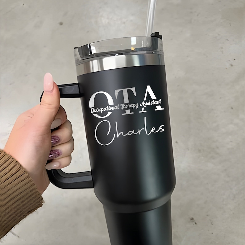 Pu&ograve; includere: Tazzina da viaggio in acciaio inossidabile nero con manico. La tazza ha un'iscrizione bianca che recita "OTA Occupational Therapy Assistant Charles".