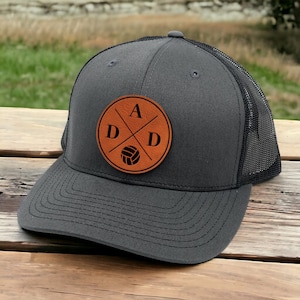 May include: A gray and black trucker hat with a brown leather patch featuring the letters "ADD" in a cross design with a volleyball in the center.