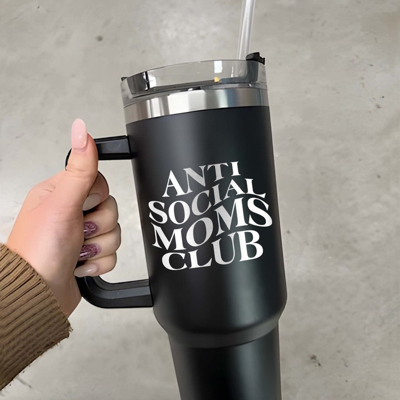 以下が含まれることがあります： 黒いステンレス製のトラベルマグ。黒いハンドルと白い文字で「ANTI SOCIAL MOMS CLUB」と書かれています。