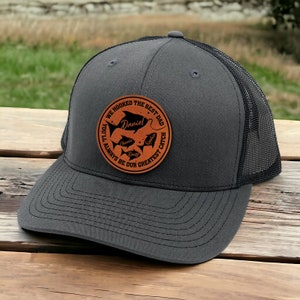 May include: A gray and black trucker hat with a brown leather patch that says "We hooked the best dad you'll always be our greatest catch" with a fish and the name "Daniel" on it.